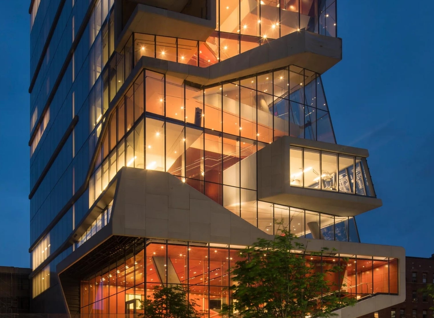 A modern, multi-story building with large glass windows is illuminated at night, showcasing its unique, angular architectural design, reminiscent of a 3D printing marvel.