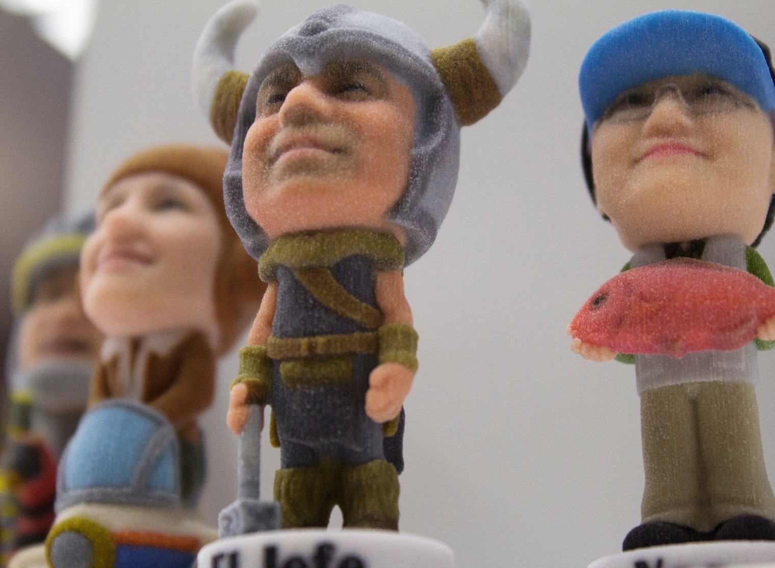 Close-up of three small, detailed figurines with exaggerated heads created using 3D printing. Left to right: a horned warrior, a person wearing a helmet, and a person with glasses holding a fish.