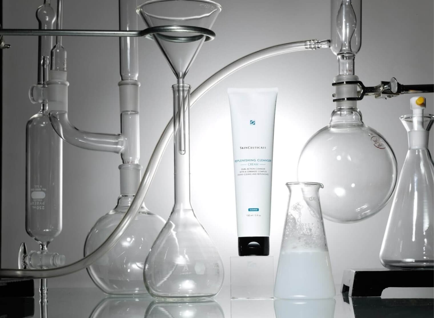 A laboratory setup with glassware, a tube of Skinceuticals Replenishing Cleanser amidst various flasks and beakers, reflecting a chemistry-themed skincare concept, is surrounded by some 3D scanning equipment in the background.