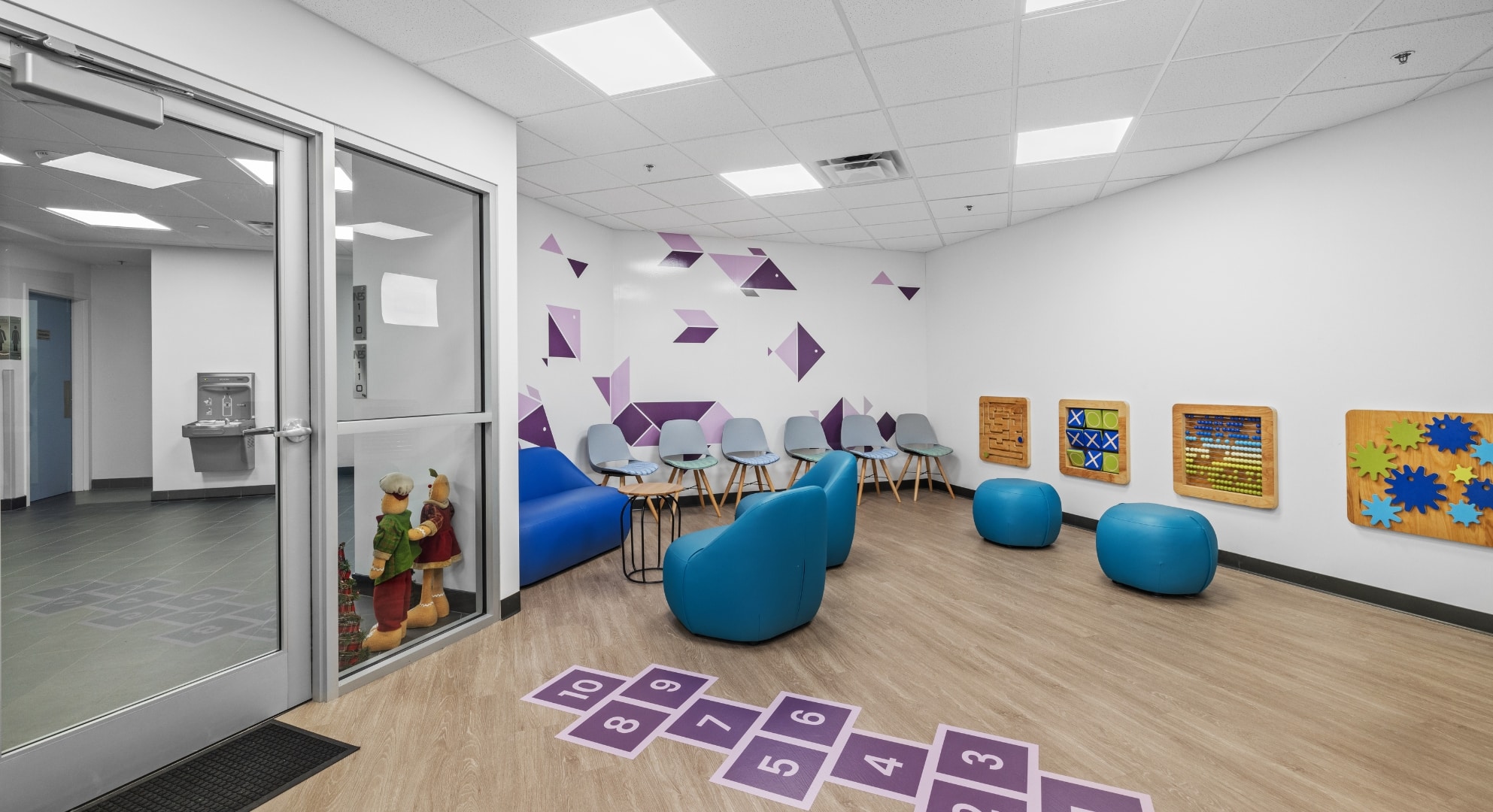 A children's waiting area with blue seating, wall-mounted interactive boards, a hopscotch floor game, and geometric wall art, accompanied by vibrant large format printing for an immersive experience.