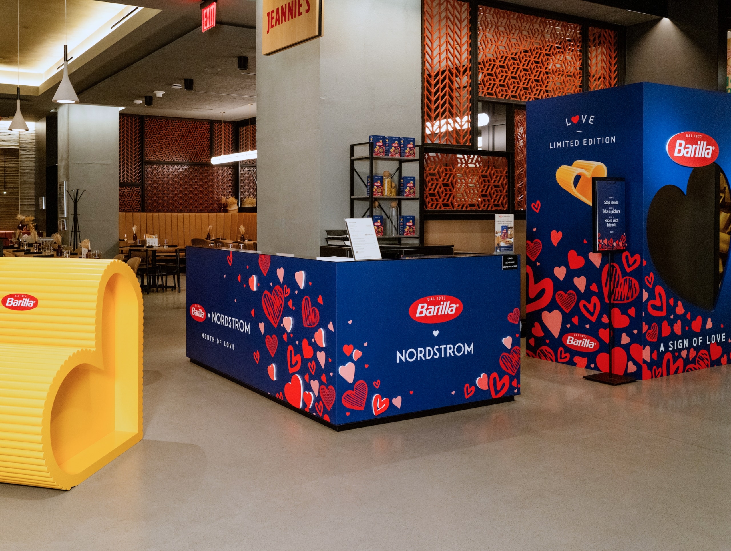 A Barilla promotional display inside a Nordstrom store, featuring a counter with branded graphics, heart designs, and product samples. In the background are dining tables and chairs, all enhanced through small format printing to create a vibrant shopping experience.