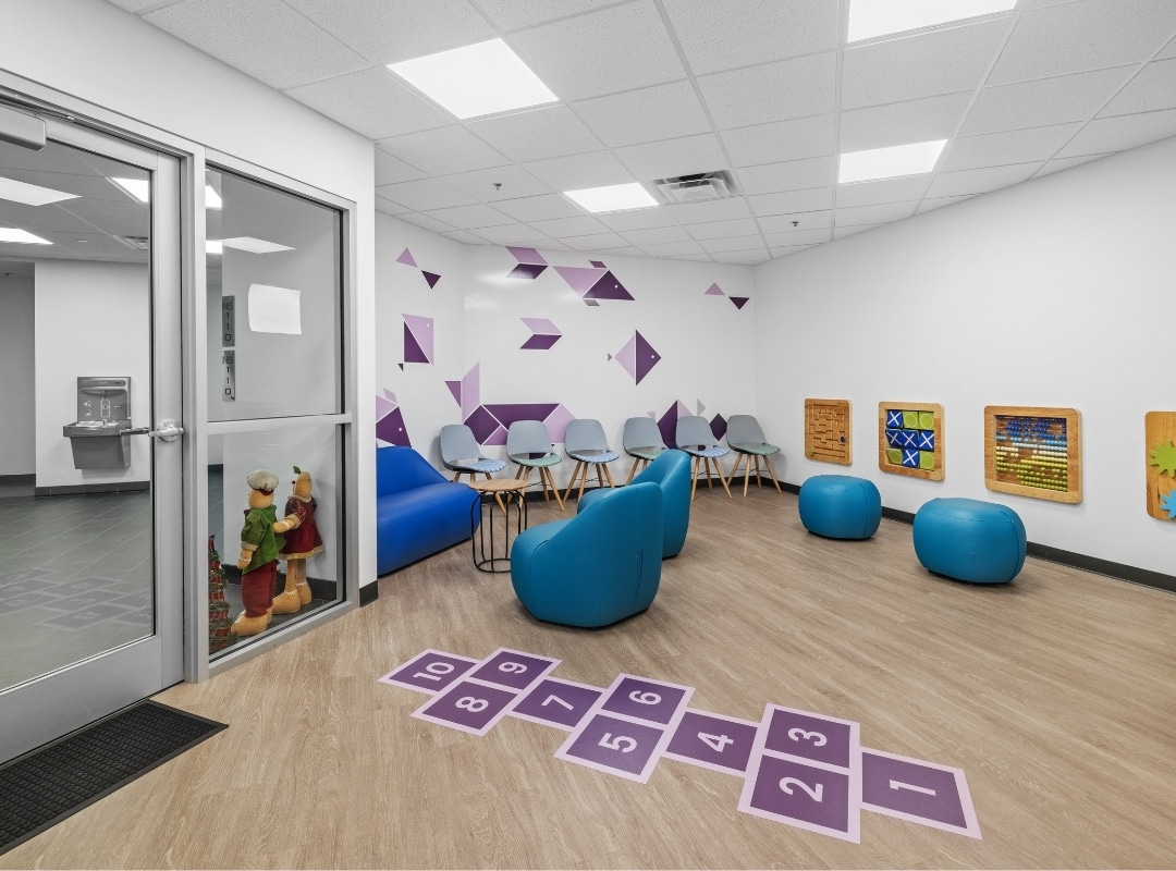 A playroom with a hopscotch mat, blue beanbags, a row of gray chairs, wall-mounted activity boards, and a glass door entrance. The walls are adorned with purple geometric shapes created using large format printing for vibrant impact.