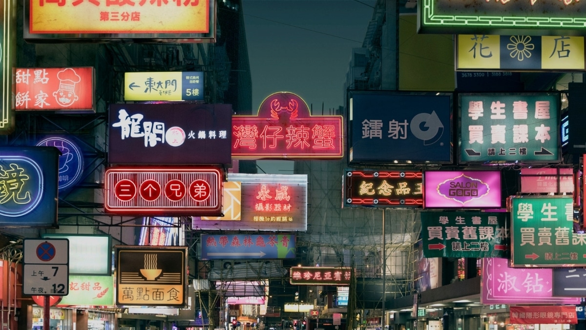 Street view with illuminated neon signs in various languages and colors, showcasing businesses offering 3D scanning services in a bustling urban area at night.