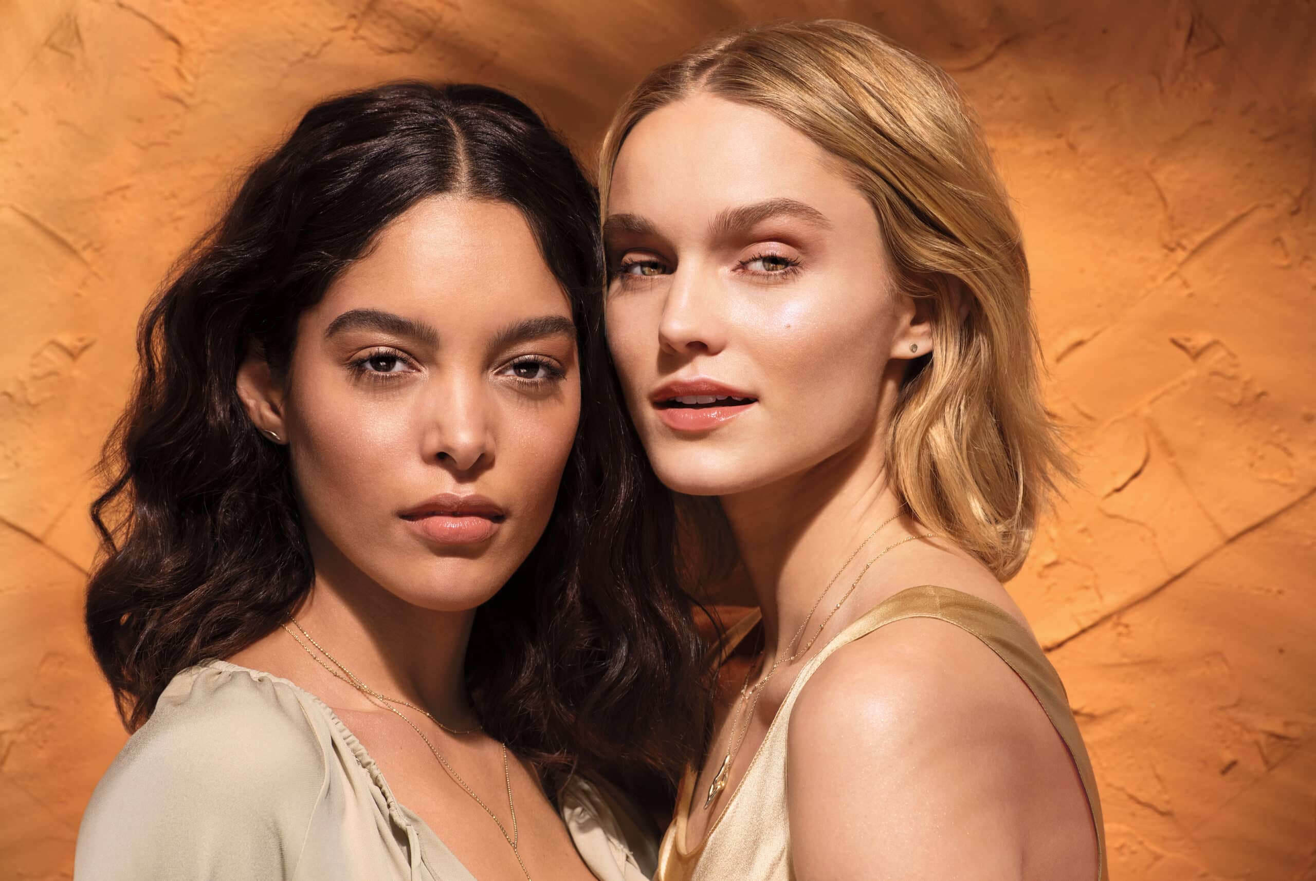 Two women with long hair stand closely against an orange textured wall, looking directly at the camera. Their light-colored tops and minimal jewelry create a harmonious visual, much like how managed IT services seamlessly integrate technology solutions.