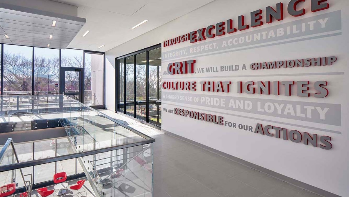 Modern office space with a wall displaying motivational words including "Grit," "Excellence," and "Culture." Outside, a glass-enclosed seating area and views of trees are visible through large windows. The design incorporates seamless integration of small format printing for personalized touches.