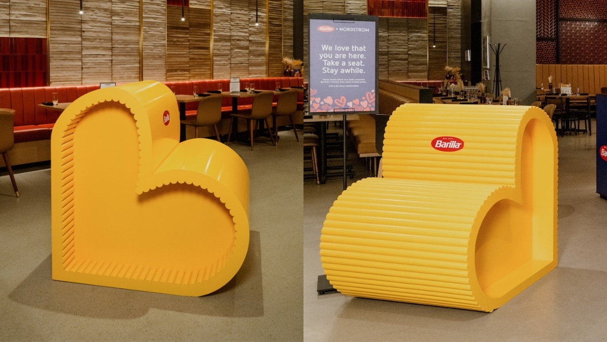 A restaurant interior featuring yellow, heart-shaped seating structures with the Barilla brand logo. A sign in the background reads, "We love that you are here. Take a seat. Stay awhile." The inviting atmosphere is complemented by vibrant large format printing adorning the walls.