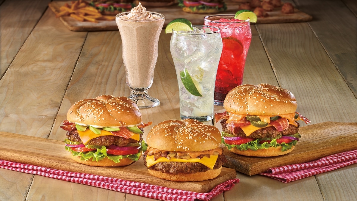 Three cheeseburgers with various toppings sit alongside a chocolate milkshake, a lime soda, and a red drink with lime on a wooden table, perfectly capturing the creativity akin to 3D printing services.