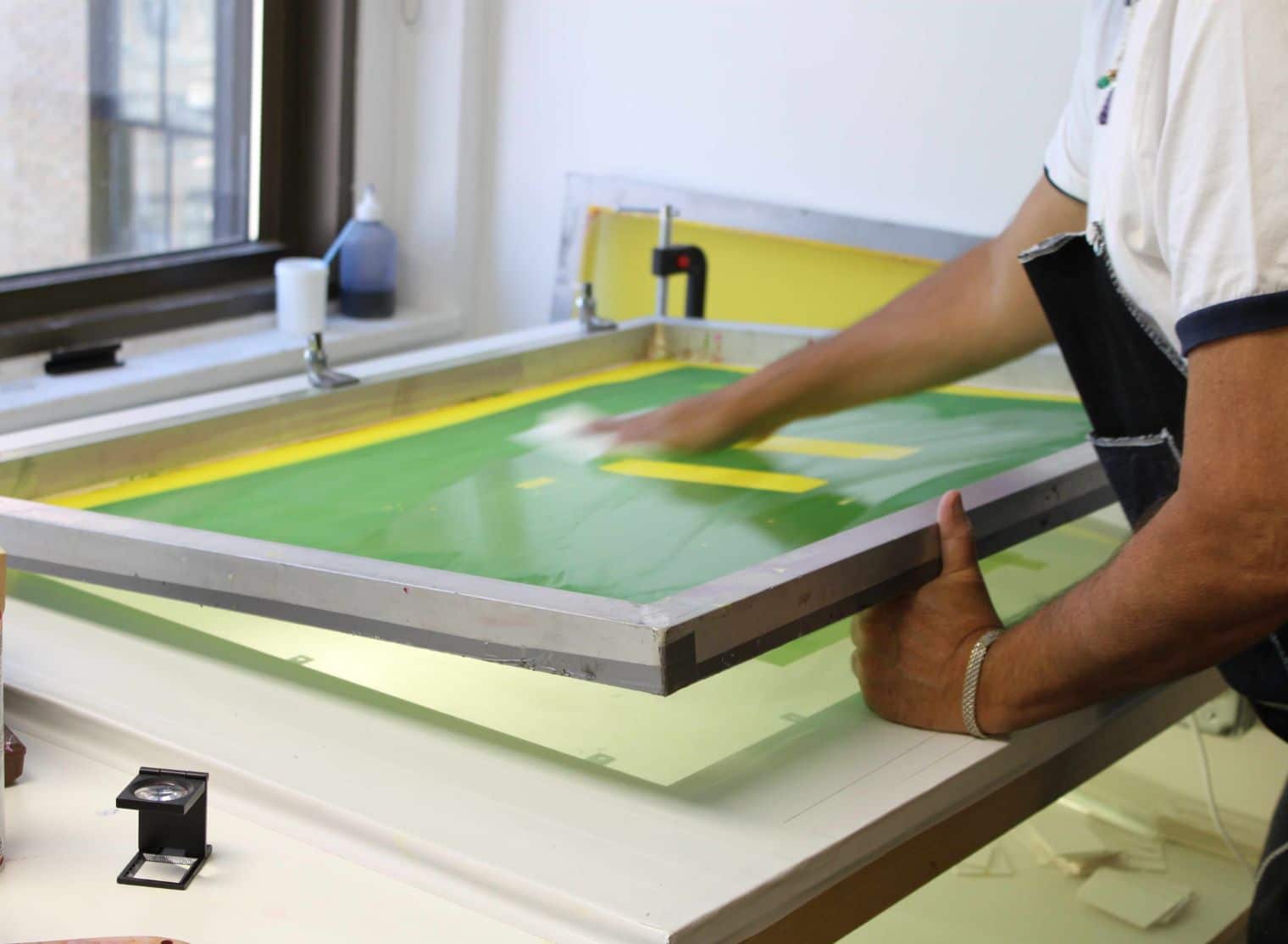 A person expertly uses a squeegee to spread ink over a screen printing frame on a table by the window, echoing precision akin to managed print services.