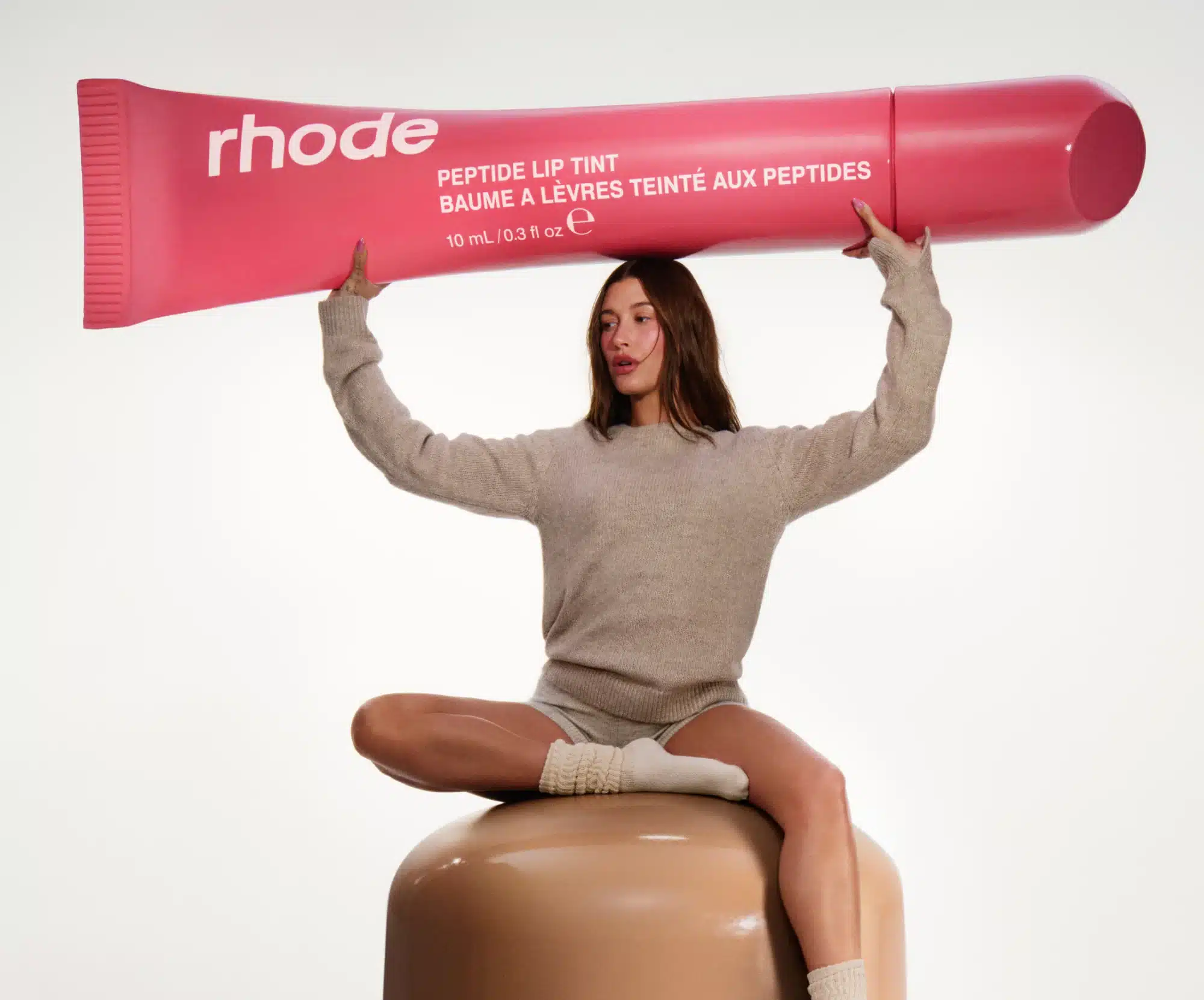 A person in a sweater and shorts sits on a large cylinder, holding an oversized pink lip tint tube labeled "rhode," showcasing the precision akin to advanced 3D scanning services.