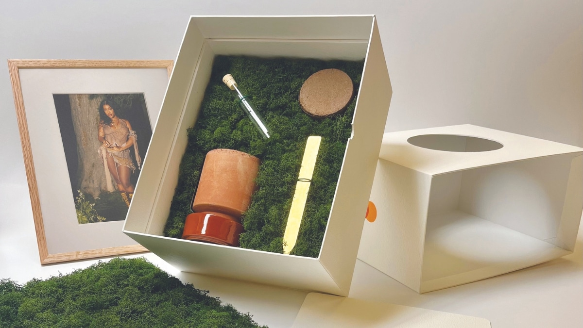 A white box containing a terracotta planter, a cork lid, a wooden test tube, and a sealed scroll, all nestled in green moss next to a framed photograph of a woman. The scene evokes an artist's studio where 3D printing and traditional crafts coexist seamlessly.