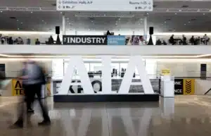 Large white "AIA" letters in the foreground with people walking past, showcasing the hub of innovation. Signs for exhibition halls and conference rooms are visible in the background, hinting at cutting-edge 3D printing services on display.