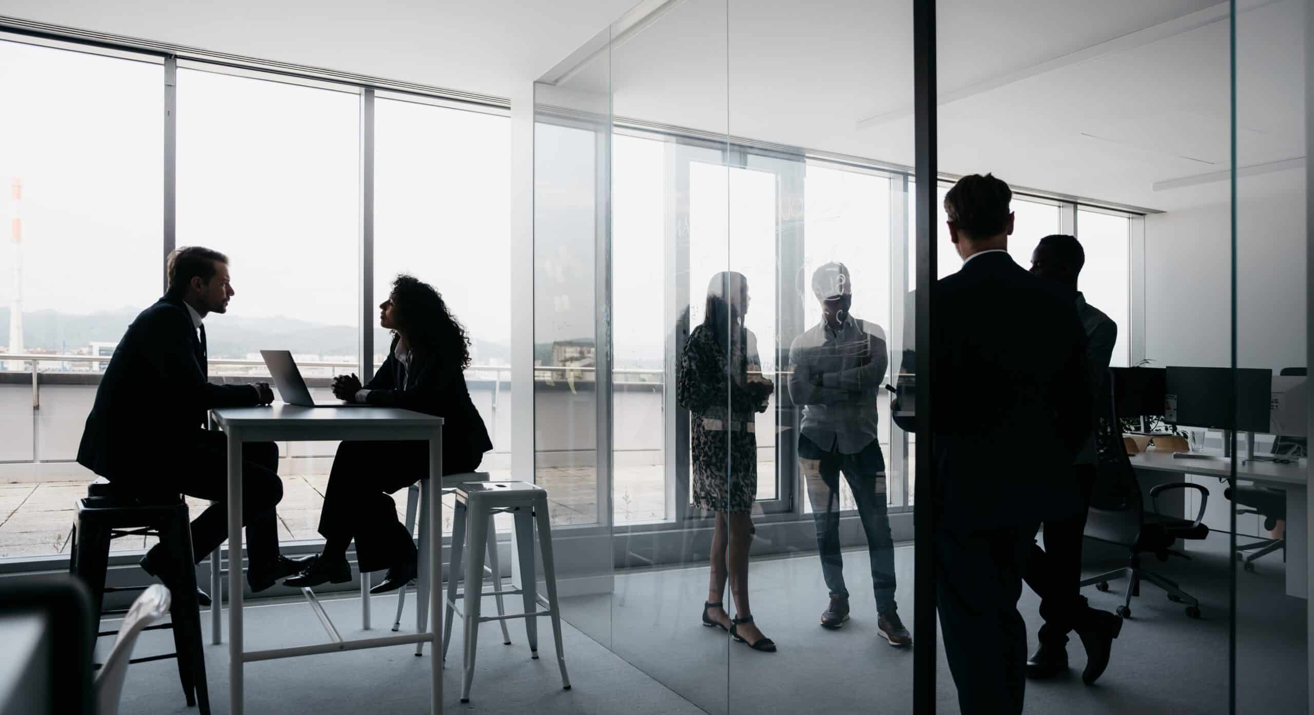 In a sleek, modern office with glass walls, a team collaborates seamlessly. Two sit at the table discussing strategies on a laptop, while others stand and converse around them, exploring innovative ideas like 3D scanning services to enhance their workflow.