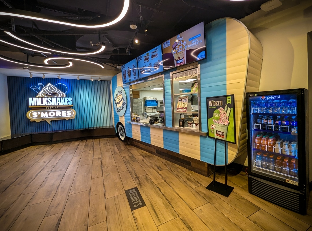 A milkshake and s’mores shop with a retro trailer-style counter, digital menu screens, a pickup window, and a cooler stocked with bottled drinks.
